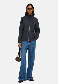 Kobieta w ciemnej bluzie zapinanej na zamek, w niebieskich szerokich dżinsach, brązowych butach, w okularach przeciwsłonecznych oraz trzymająca małą, okrągłą brązową torebkę.