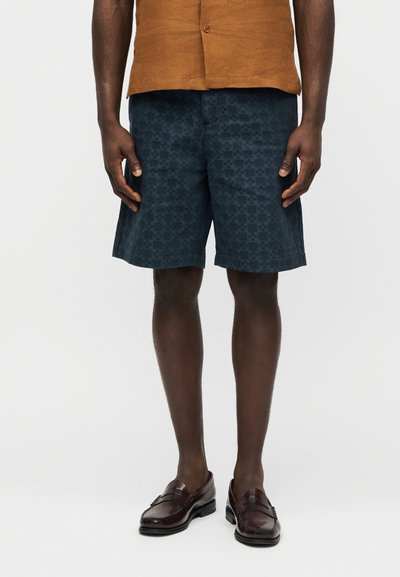 Άνδρας που φοράει σκούρα σορτς με σχέδια, καφέ μοκασίνια και καφέ πουκάμισο με κοντά μανίκια και κουμπιά, στέκεται μπροστά σε απλό φόντο.