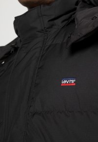 Svart puffjacka med hög krage, som har ett litet logobadge i röd, vit och blå på bröstet och en slät, strukturerad tyg.