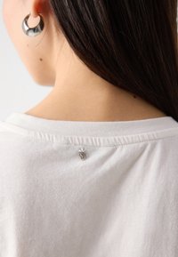 Tricou alb din bumbac, cu decolteu rotund, având un mic detaliu argintiu la decolteul din spate și o textură netedă, moale.