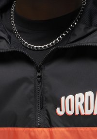 Giacca nera e arancione con zip, dal finish lucido, caratterizzata da un grande logo "JORDAN" e da una collana in argento visibile.