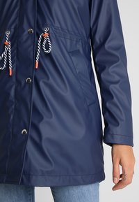 Personne portant un imperméable bleu avec cordons en corde et boutons-pression métalliques, main gauche visible, jean bleu clair en dessous.