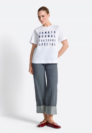 Woman wearing white T-shirt with French text and blue striped wide-leg pants, standing with one hand behind back and red ballet flats.