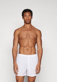 Hvite bomullshorts med elastisk liv, løs passform og et enkelt design, båret av en stående mannelige modell mot en nøytral bakgrunn.