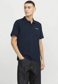 Marineblå polo T-shirt med korte ærmer, ribbet krave og et diskret logo på brystet. Båret med mørke denim jeans.