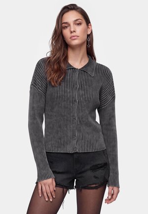 Ung kvinde med langt brunt hår iført en grå ribbet knap-sweater, sorte slidte shorts og stjerneformede hængende øreringe.