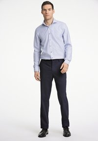 Lichtblauw gemêleerd overhemd met lange mouwen, gecombineerd met donkerblauwe broek en zwarte nette schoenen. Detail van een button-down kraag.