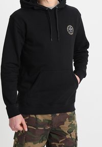 Felpa nera con cappuccio regolabile, tasca a marsupio e patch rotondo. Tessuto strutturato, indossata sopra pantaloni cargo mimetici.