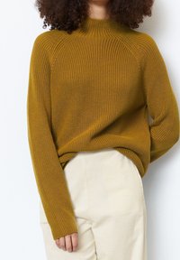 Femme portant un pull en maille jaune moutarde et un pantalon en velours côtelé crème, avec des cheveux bouclés visibles des épaules vers le haut.