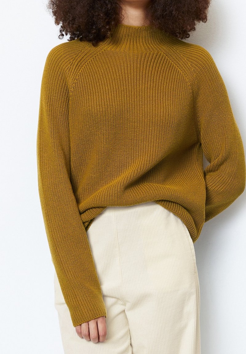 Femme portant un pull en maille jaune moutarde et un pantalon en velours côtelé crème, avec des cheveux bouclés visibles des épaules vers le haut.