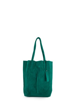 Zielona torba typu tote zamszowa z długimi uchwytami i zawieszką ze skóry, stojąca pionowo na białym tle.