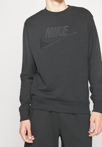 Person som bär en svart Nike-sweatshirt och matchande svarta byxor, står med avslappnade händer längs sidorna.