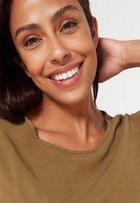 Brązowa koszula z krótkim rękawem i okrągłym dekoltem; modelka uśmiecha się, ukazując proste białe zęby i gładką, opaloną skórę.