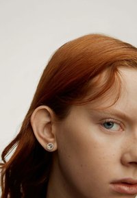 Pendiente de plata en forma de círculo, con una piedra central rodeada de pequeños cristales. Lleva puesto un modelo con cabello rojo.