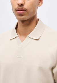Άνδρας που φοράει μπεζ πόλο πουκάμισο με υφή, με ριμπ λαιμόκοψη και μικρό οβάλ λογότυπο στο στήθος, εμφανιζόμενος από τον λαιμό έως τη μέση του κορμού.