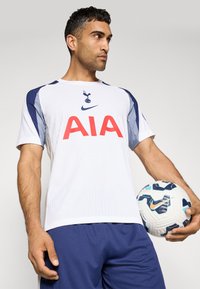 Hvid sports T-shirt med blå accenter, AIA-logo og et lille emblem på brystet, parret med marineblå shorts og holder en mønsterfodbold.