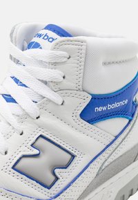 Gros plan d'une basket New Balance blanche et bleue montrant les lacets, les coutures, le logo sur le côté et le col rembourré autour de la cheville.