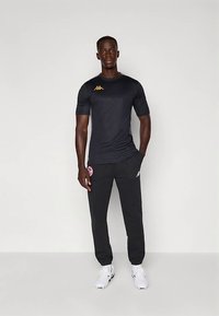 Crna sportska majica kratkih rukava s zlatnim logom, u kombinaciji s crnim hlačama na kojima se nalazi crveni logo, i bijelim tenisicama.