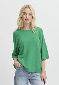 Grüner gestrickter Pullover mit rundem Ausschnitt, 3/4-Ärmeln und geripptem Saum. Getragen über hellblauen Jeans. Weiche Textur, lässige Passform.