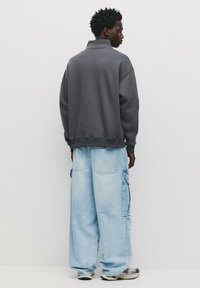 Grauer Pullover mit hohem Kragen, kombiniert mit hellblauen Cargo-Hosen, die seitliche Taschen und eine lockere Passform aufweisen. Beide Teile haben eine strukturierte Oberfläche.