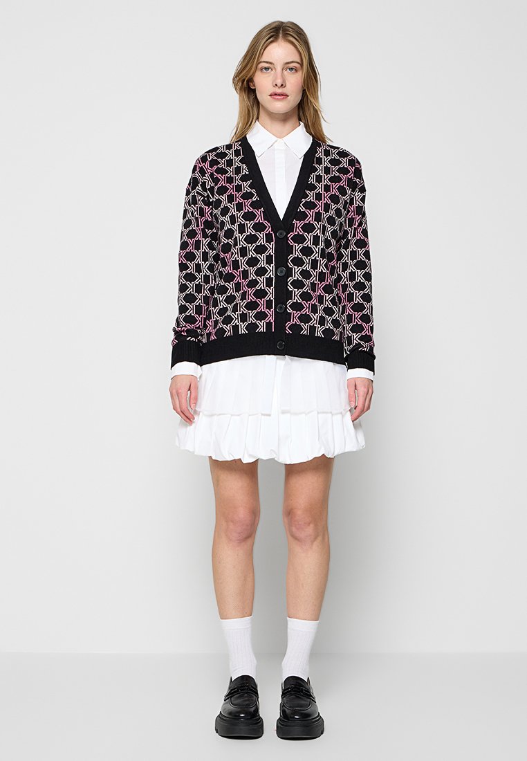 Schwarzer Cardigan mit geometrischem Muster in Pink und Creme, Knopfleiste, V-Ausschnitt, gerippte Bündchen, getragen über einem weißen Rüschenkleid und schwarzen Schuhen.