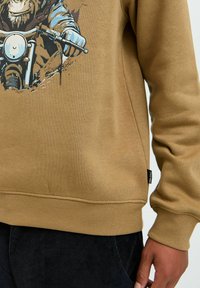 Brun sweatshirt med grafik af en chimpanse på en motorcykel. Ribbet manchetter og kant, blødt stof med en glat tekstur.