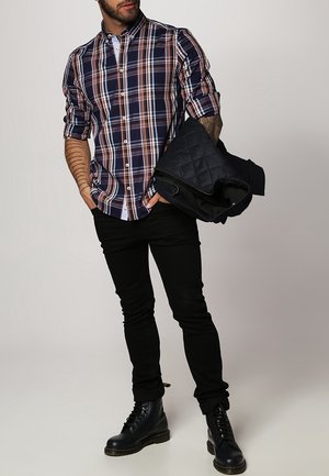 Muž v zrolované kárované košili, černých džínách a černých botách, držící v pravé paži složenou černou prošívanou bundu.
