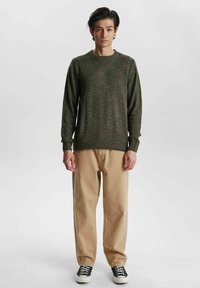 Maglione verde lavorato a maglia con scollatura rotonda, polsini e orlo a coste, abbinato a pantaloni beige chiaro e sneakers nere.