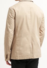 Giacca leggera beige con una trama liscia, taglio dritto e un'unica fessura posteriore. Presenta polsini con bottoni e un design minimalista.