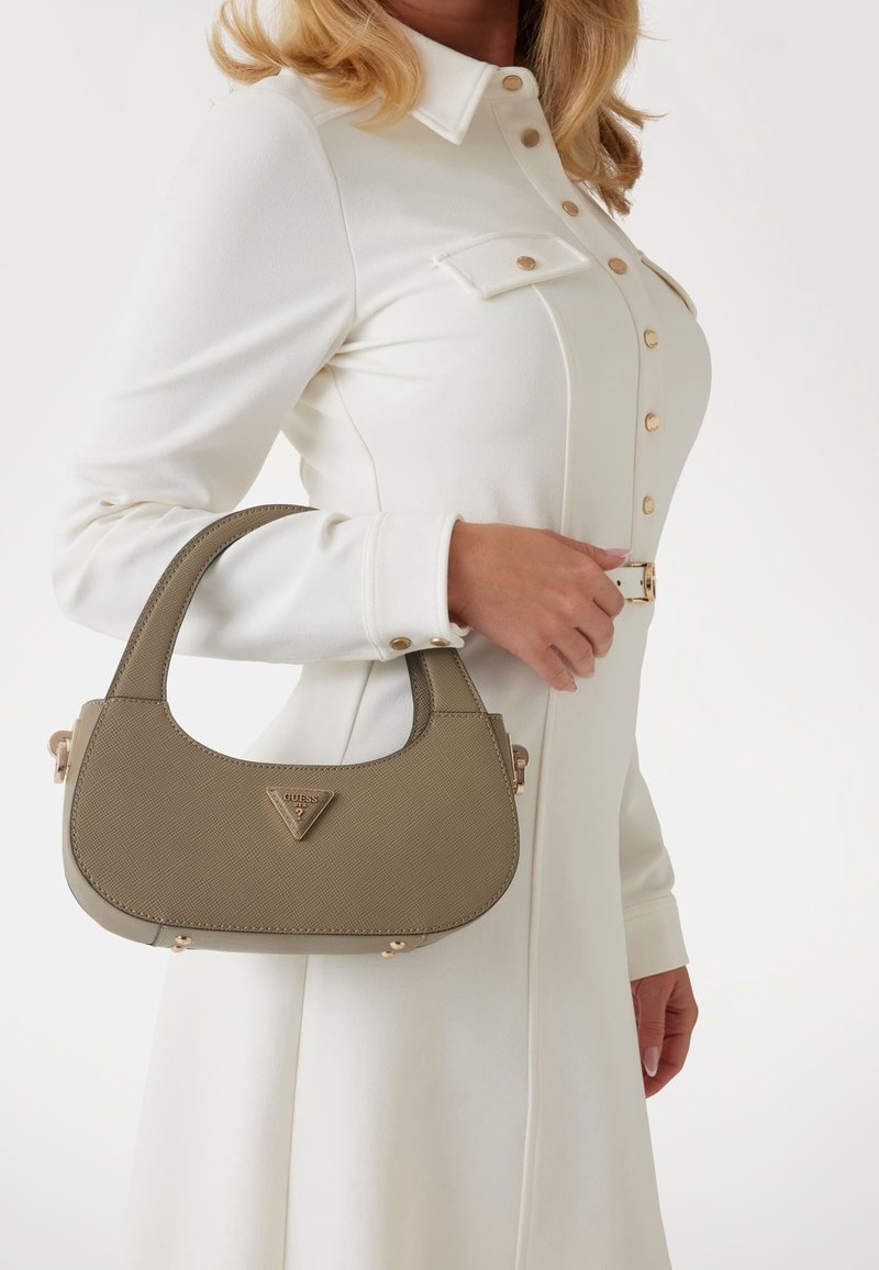 Femme en robe blanche boutonnée tenant un sac à main beige Guess avec des poignées courtes sur son bras, sur un fond uni.