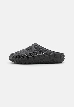 Zueco negro sin cordones con patrón geométrico de diamantes y ranuras de ventilación laterales, presentado sobre un fondo blanco.
