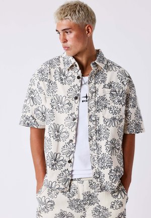 Jeune homme aux cheveux blonds courts portant une chemise à manches courtes couleur crème et un short assorti avec des motifs floraux abstraits noirs.