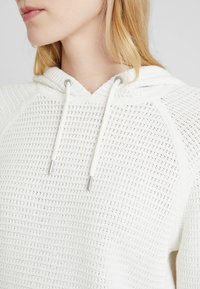 Biała dziergana bluza z kapturem, ozdobiona teksturowanym wzorem, z kapturem na sznurku z metalowymi akcentami oraz rękawami typu raglan.