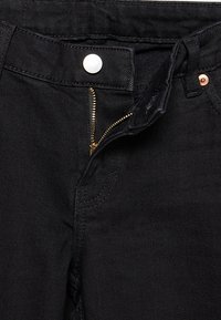 Gros plan d'un jean en denim noir avec une fermeture éclair dorée ouverte, un bouton argenté et des coutures visibles autour de la ceinture et des poches.