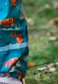 Regnjakke i teal med tegneserie-køer i forskellige positioner, med en glat tekstur og reflekterende stribe nær kanten.