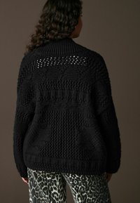 Pulover negru tricotat cu un model texturat, având un guler înalt și detalii de lucru deschis pe spate. Design lejer.