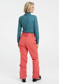 Maglia a maniche lunghe color teal abbinata a pantaloni da sci color corallo, caratterizzati da una cintura testurizzata e gambe con zip, progettati per attività sportive invernali.