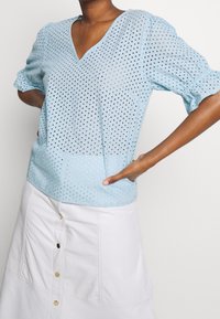 Jasnoniebieska bluzka z krótkim rękawem i dekoltem w kształcie litery V, z perforowanym wzorem i teksturowanym materiałem, zestawiona z białą spódnicą na guziki.