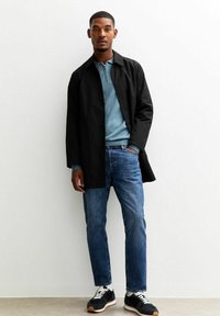 Zwarte lange jas over een lichtblauwe zip-up sweater, gecombineerd met blauwe jeans en zwarte sneakers met witte veters en tan zolen.