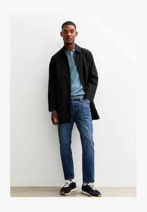 Un long manteau noir porté sur un pull zippé bleu clair, associé à un jean bleu et des baskets noires avec des lacets blancs et des semelles beige.