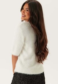 Donna con lunghi capelli scuri e mossi, sorridente e che guarda oltre la spalla, indossa un maglione bianco corto e peloso con maniche corte e una gonna nera con trama.