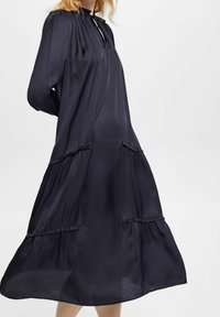 Mörkblå klänning i silkesliknande tyg, med veckad design, långa ärmar, nyckelhålsurringning och volangdetaljer på kjolen.