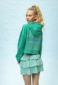 Groene cropped hoodie met capuchon, voorzien van een herhaald tekstontwerp op de achterkant. Gecombineerd met een gelaagde groene gestippelde rok.