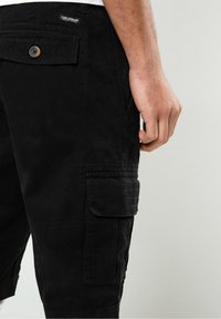Bermudas negras de carga fabricadas con un tejido duradero, que cuentan con bolsillos laterales, un color sólido y un acabado texturizado. Botones y detalles de costura visibles.