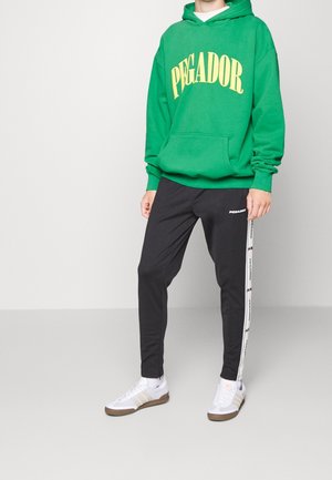 Persoon in een groene hoodie met "PEGADOR" in het geel, zwarte broek met logo en witte sneakers, staande tegen een effen achtergrond.
