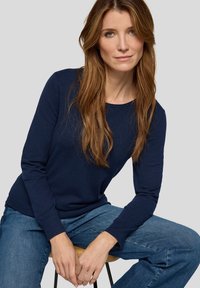 Marineblauer Strickpullover mit rundem Ausschnitt und gerippten Bündchen, kombiniert mit hellblauen Jeanshosen. Die Textur wirkt weich und angenehm.