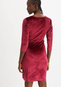 Abito rosso velluto con un design aderente, maniche a tre quarti e una delicata spruzzata di dettagli glitterati su tutta la stoffa.
