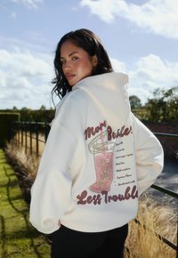 Vrouw met donker haar die een witte hoodie draagt met een bubble tea graphic en de tekst "Meer Bubbles Minder Probleem," staande buiten bij een reling.