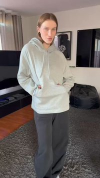 Light gray hoodie with a kangaroo pocket, drawstrings, and relaxed fit, paired with dark wide-leg pants. Soft fabric and casual design.