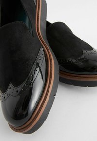 Scarpe slip-on nere con punta in vernice lucida, tomaia in suede e suola in gomma marrone. Presentano dettagli brogue lungo i bordi.